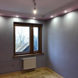 LASER - Wykończenia Wnętrz - Pomalowane na fioletowo wnętrze pokoju z drewnianą podłogą w jodełkę, brązowe okno z widokiem na zewnątrz, biały grzejnik i sufit z oświetleniem punktowym.