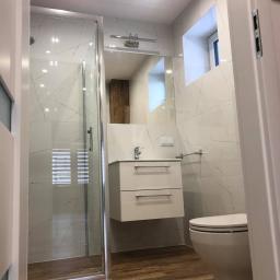 LASER - Wykończenia Wnętrz - Nowoczesna łazienka z kabiną prysznicową, białymi płytkami imitującymi marmur, wiszącą szafką z umywalką i drewnopodobną podłogą, widok zza otwartych drzwi.