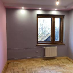 LASER - Wykończenia Wnętrz - Pokój po odświeżeniu: ściany pomalowane na liliowo i różowo, drewniana podłoga parkietowa, okno w drewnianej ramie, biały grzejnik, widoczne punkty oświetleniowe w suficie.