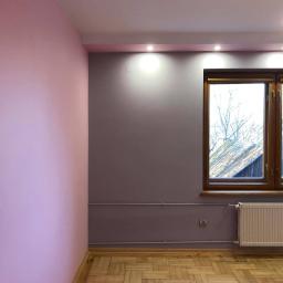 LASER - Wykończenia Wnętrz - Pomieszczenie po odświeżeniu: różowa ściana kontrastuje z szarą, drewniana podłoga w jodełkę, okno z widokiem na drzewa, biały grzejnik.