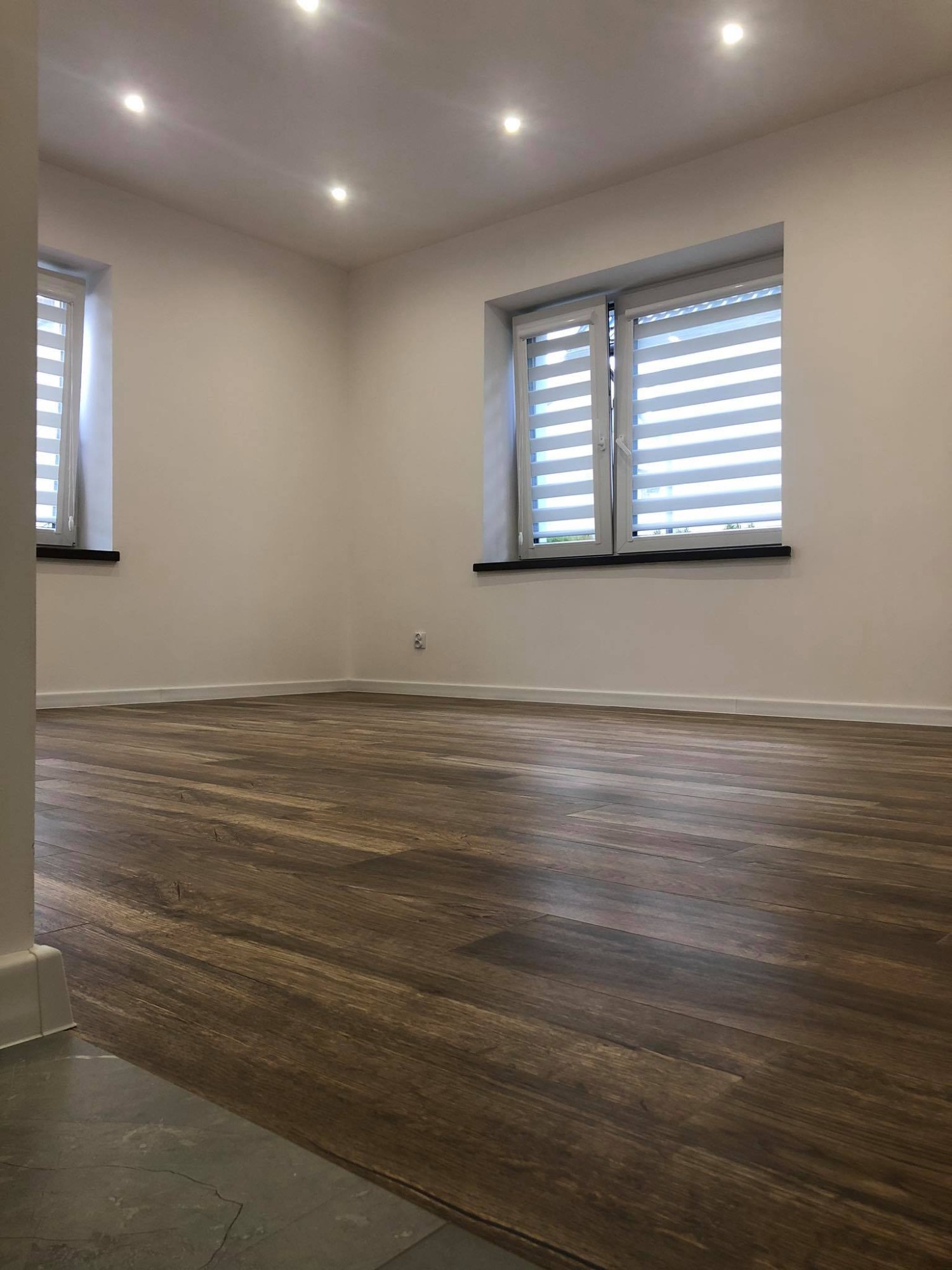 Jasne, nowo wykończone wnętrze z drewnianą podłogą, białymi ścianami, dwoma oknami z żaluzjami dzień-noc i oświetleniem punktowym w suficie.
