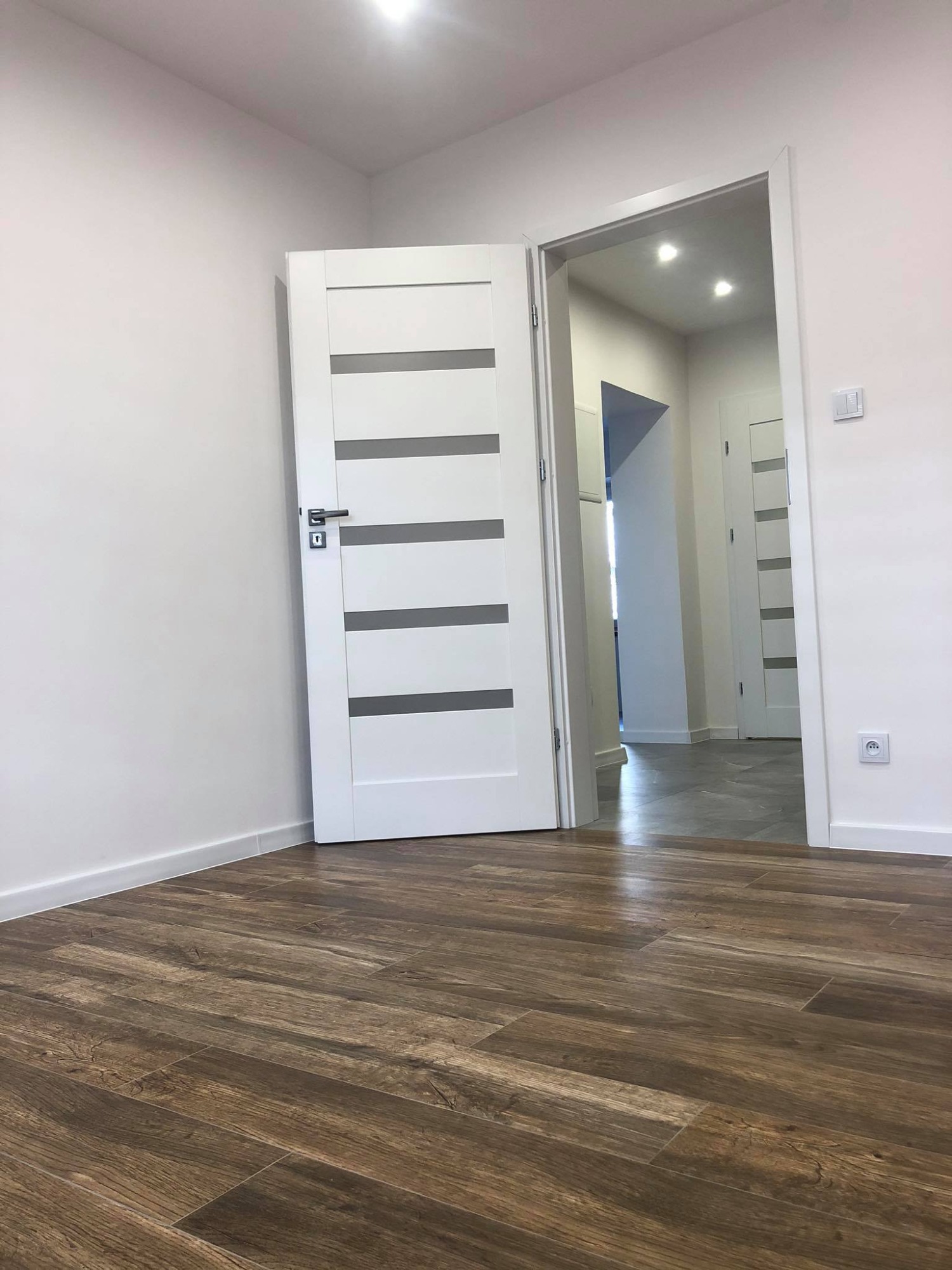 Nowoczesne wnętrze z drewnopodobną podłogą, białe ściany i białe drzwi wewnętrzne z poziomymi szarymi paskami, widok na korytarz z oświetleniem punktowym.