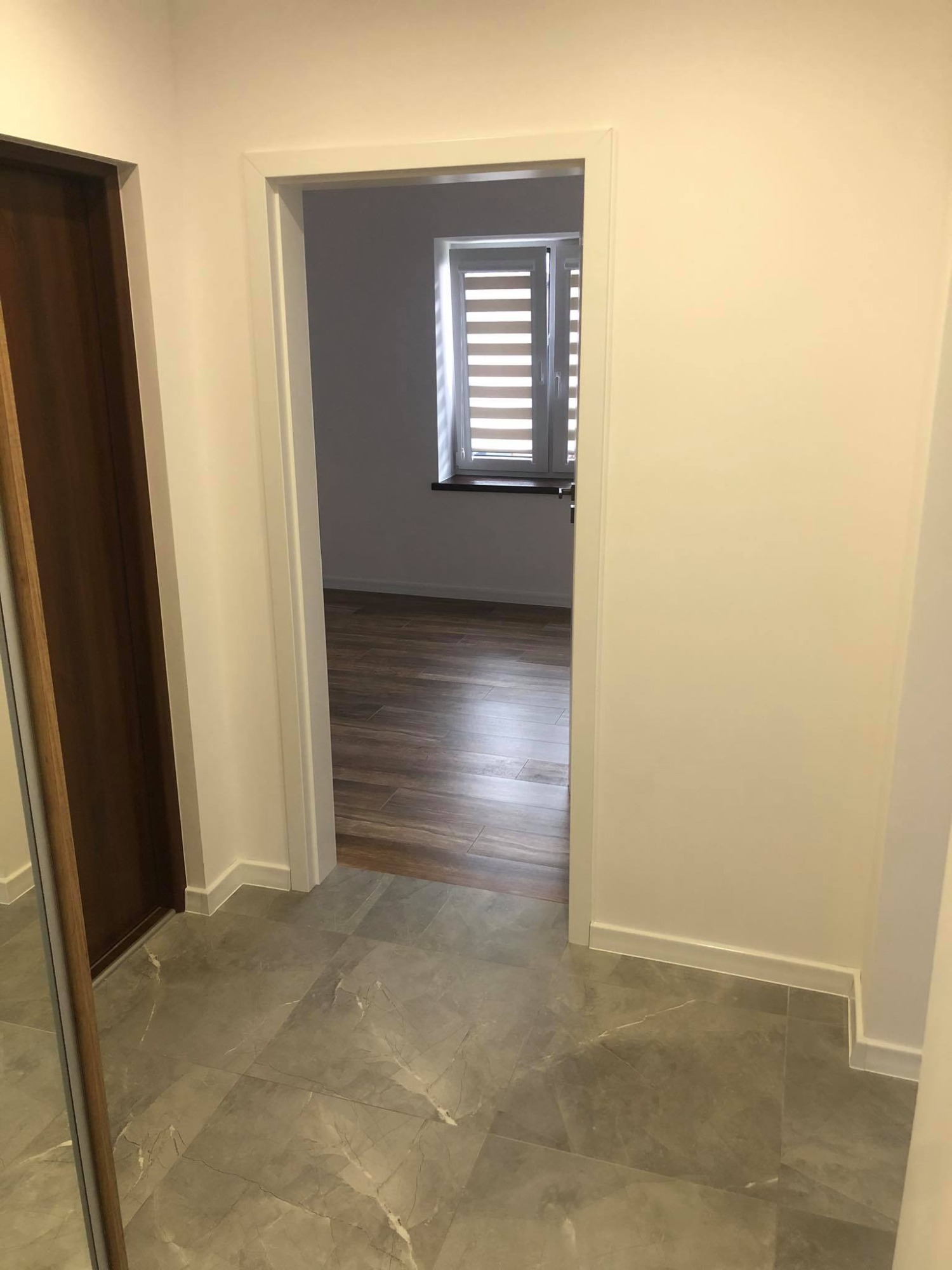 Wykończony korytarz z szarą marmurkową podłogą, widok na sąsiedni pokój z drewnianą podłogą i oknem z żaluzjami.