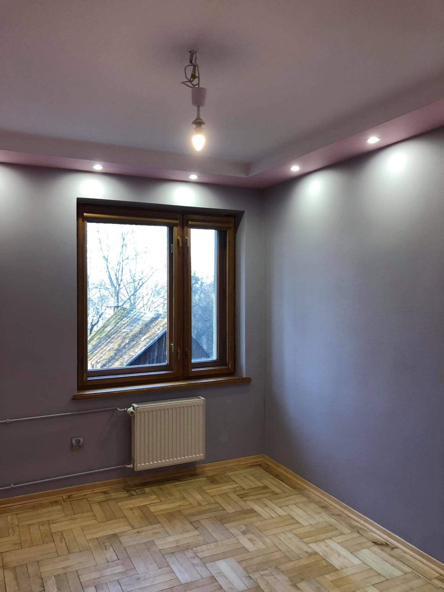 Pomalowane na fioletowo wnętrze pokoju z drewnianą podłogą w jodełkę, brązowe okno z widokiem na zewnątrz, biały grzejnik i sufit z oświetleniem punktowym.