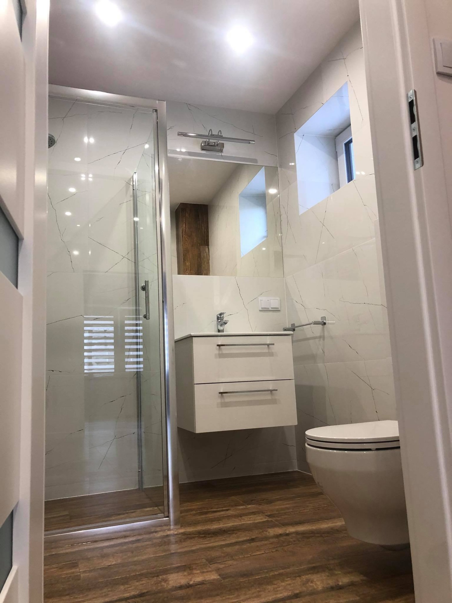 Nowoczesna łazienka z kabiną prysznicową, białymi płytkami imitującymi marmur, wiszącą szafką z umywalką i drewnopodobną podłogą, widok zza otwartych drzwi.