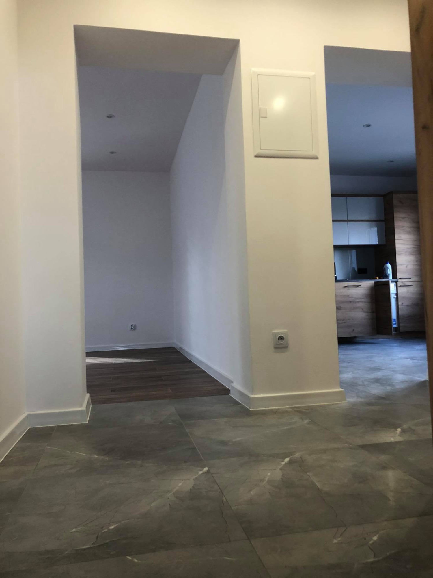 Wnętrze mieszkania po remoncie, widok z korytarza na nową podłogę z szarych płytek imitujących marmur, fragment kuchni z drewnianymi szafkami w tle, białe ściany z gładzią i listwami przypodłogowymi.