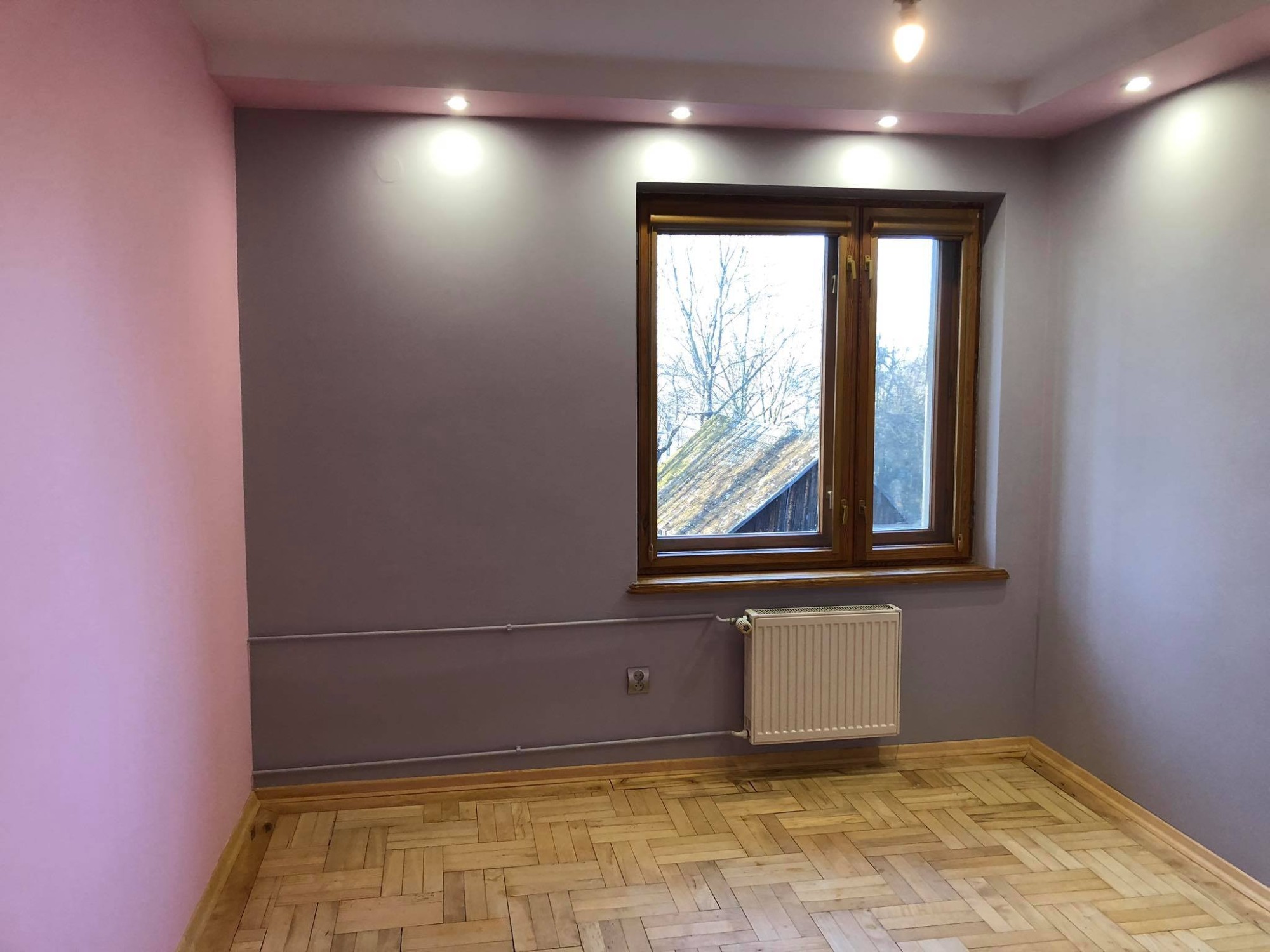 Pokój po odświeżeniu: ściany pomalowane na liliowo i różowo, drewniana podłoga parkietowa, okno w drewnianej ramie, biały grzejnik, widoczne punkty oświetleniowe w suficie.