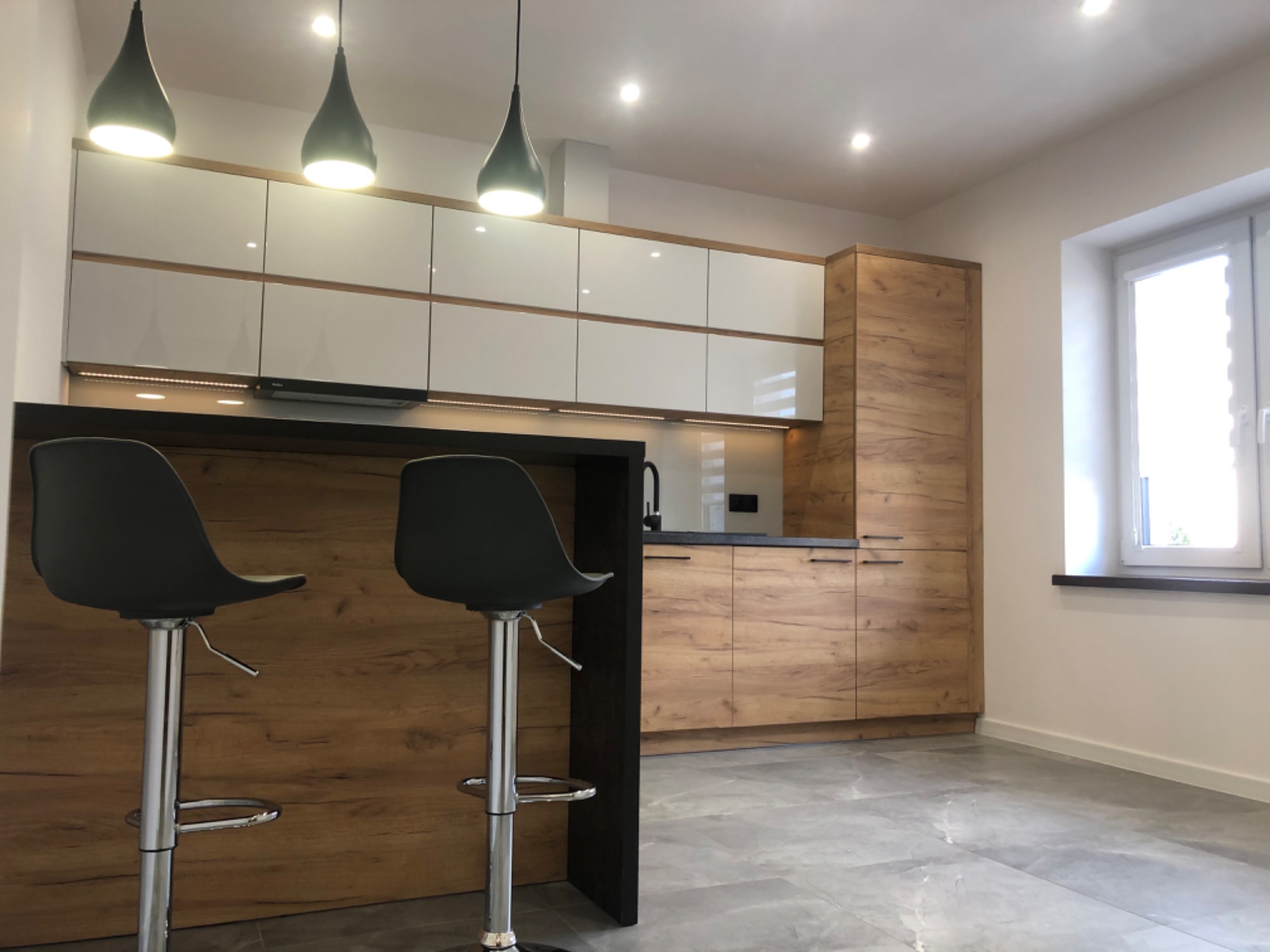 Nowoczesna kuchnia z białymi szafkami górnymi i drewnopodobnymi dolnymi, czarnym blatem, szarymi płytkami podłogowymi oraz dwoma czarnymi hokerami przy wyspie.