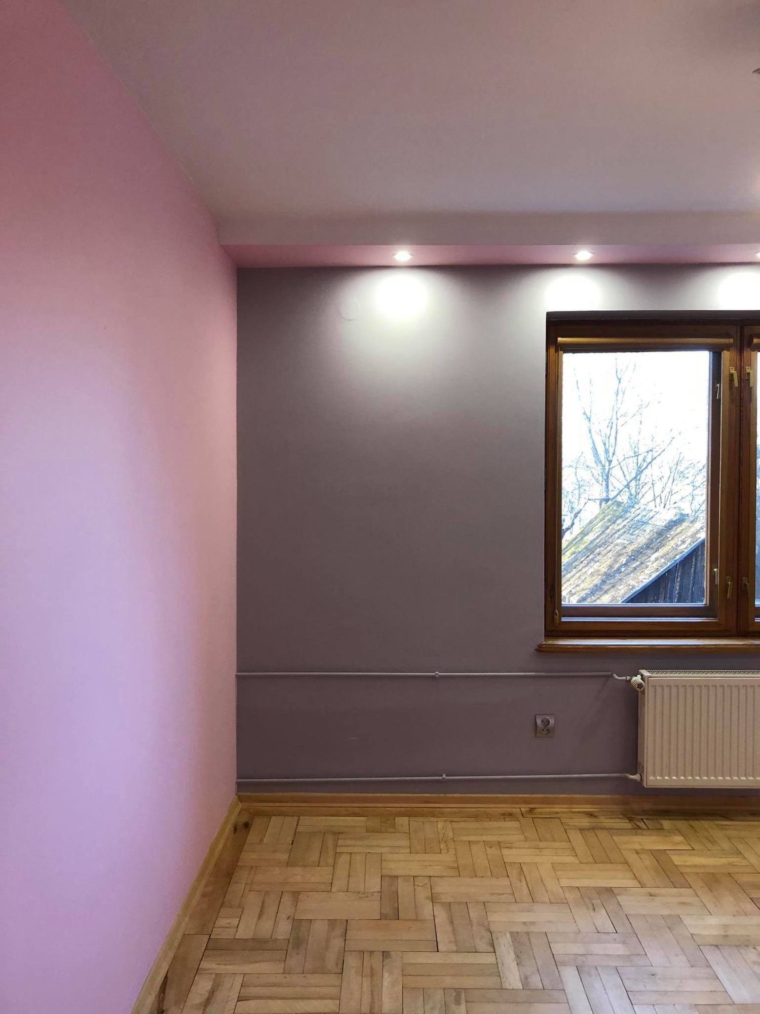 Pomieszczenie po odświeżeniu: różowa ściana kontrastuje z szarą, drewniana podłoga w jodełkę, okno z widokiem na drzewa, biały grzejnik.