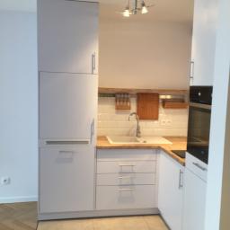 Master Format - Nowoczesna kuchnia w odcieniach bieli i szarości, z drewnianym blatem, białymi płytkami na ścianie, lampą sufitową z kilkoma reflektorami i podłogą z beżowych płytek.