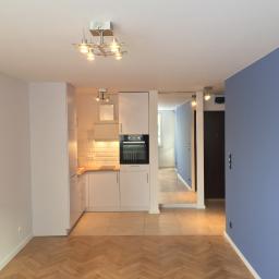 Master Format - Nowoczesna, biała kuchnia na wymiar z drewnianym blatem i białymi płytkami na ścianie, widoczna z perspektywy pokoju z parkietem w jodełkę i niebieską ścianą.