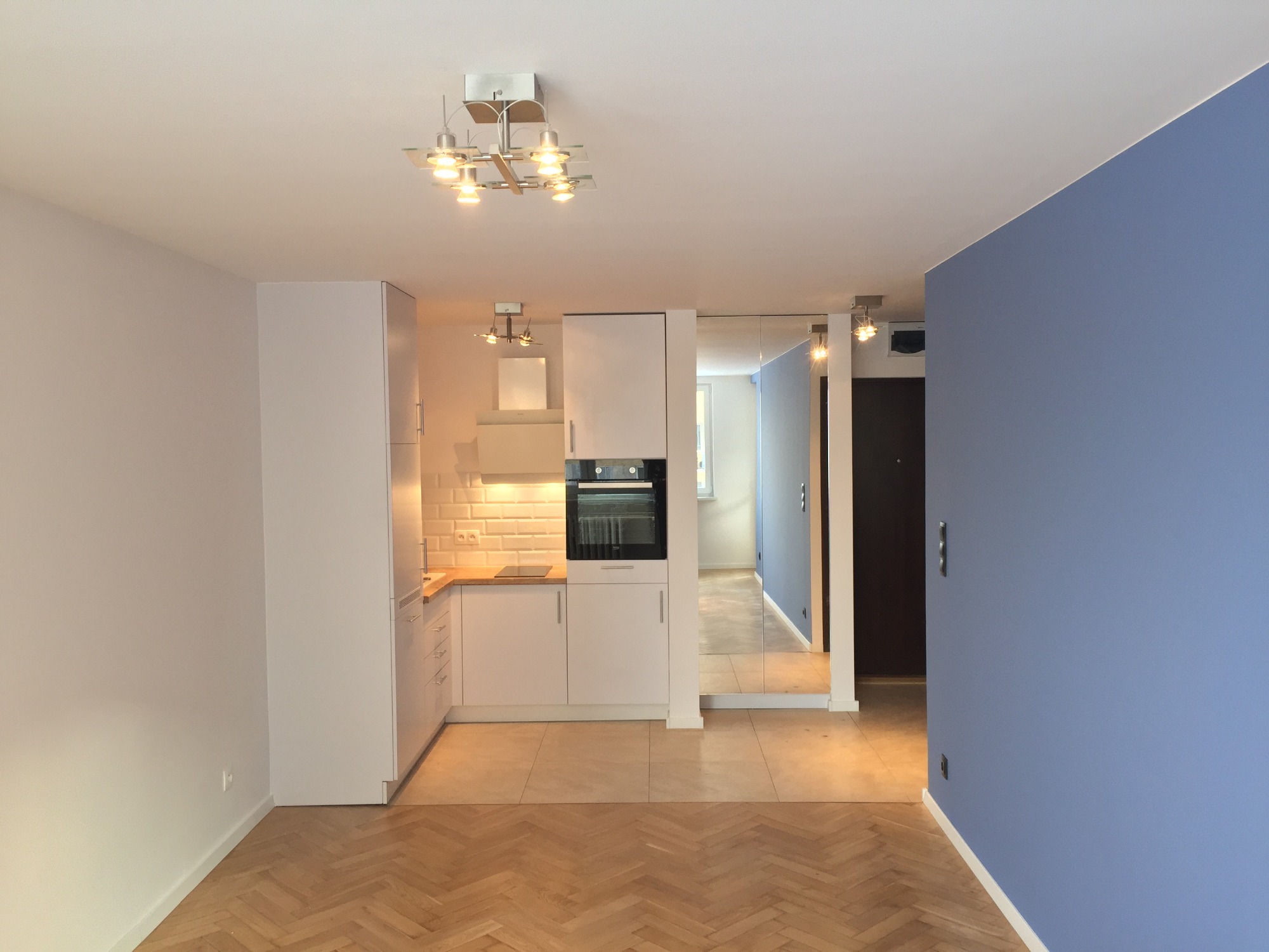 Nowoczesna, biała kuchnia na wymiar z drewnianym blatem i białymi płytkami na ścianie, widoczna z perspektywy pokoju z parkietem w jodełkę i niebieską ścianą.