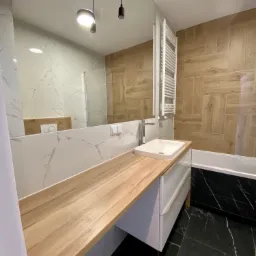 Nowoczesna łazienka z drewnianym blatem, umywalką nablatową, marmurowymi płytkami na ścianie i podłodze, oraz grzejnikiem drabinkowym na ścianie wyłożonej płytkami imitującymi drewno.