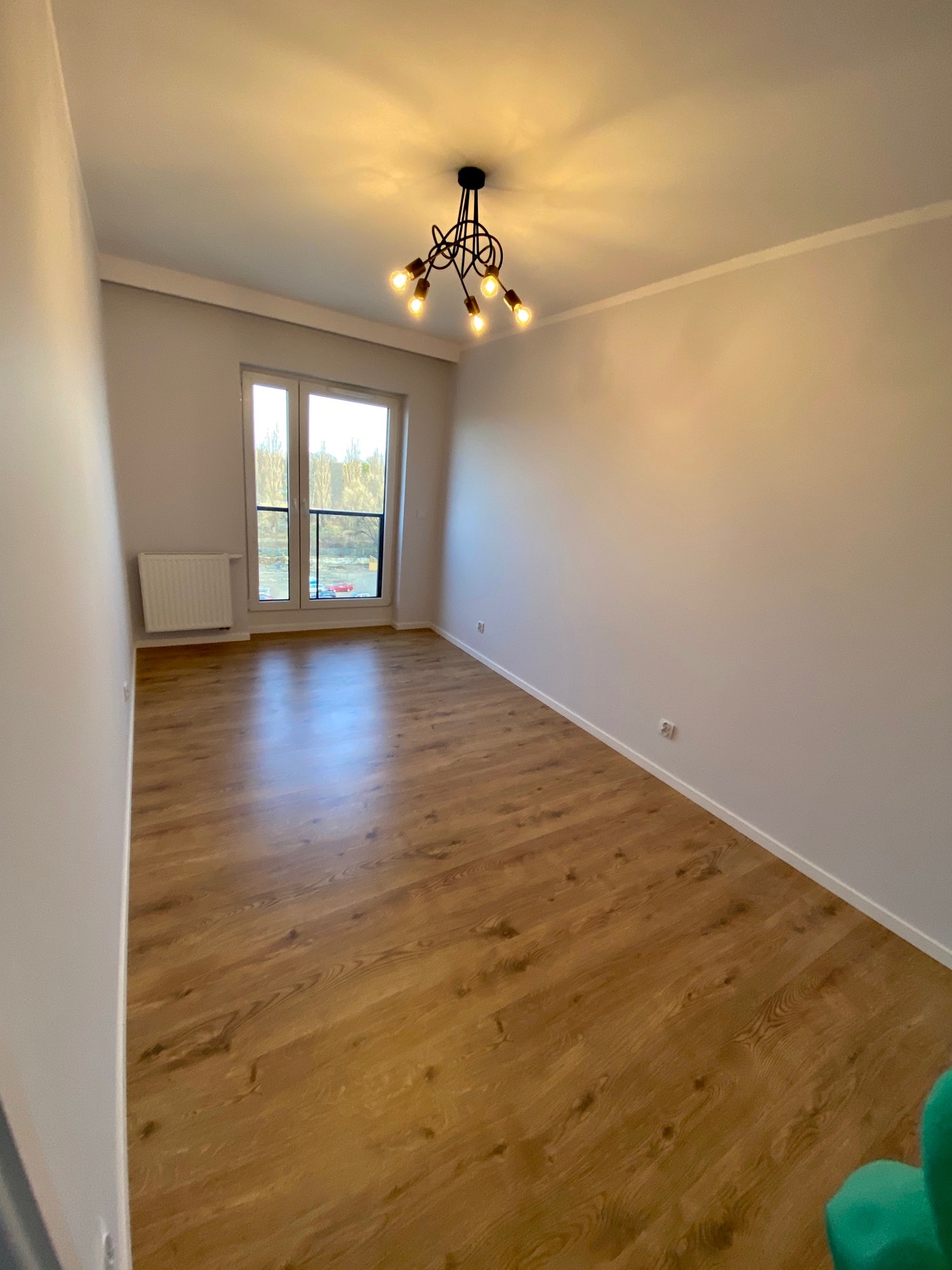 Pusty pokój po remoncie z panelami podłogowymi w odcieniach brązu, szare ściany, okno z widokiem na zewnątrz i nowoczesna lampa sufitowa z kilkoma żarówkami.