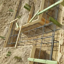 Zbrojenie fundamentu w wykopie, drewniane szalunki z zielonymi rozpórkami w piaszczystej ziemi.