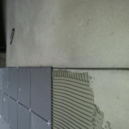 Wizner Usługi Budowlane Krystian Jurkowski - Układanie ciemnoszarych płytek na ścianie z użyciem krzyżyków dystansowych, widoczna warstwa kleju z charakterystycznym wzorem i nożyk do cięcia płytek.