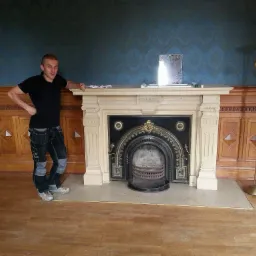 Mężczyzna w roboczych spodniach opiera się o ozdobny, odnowiony kominek z czarnym wkładem w pokoju z drewnianą boazerią i wzorzystą, niebieską tapetą.