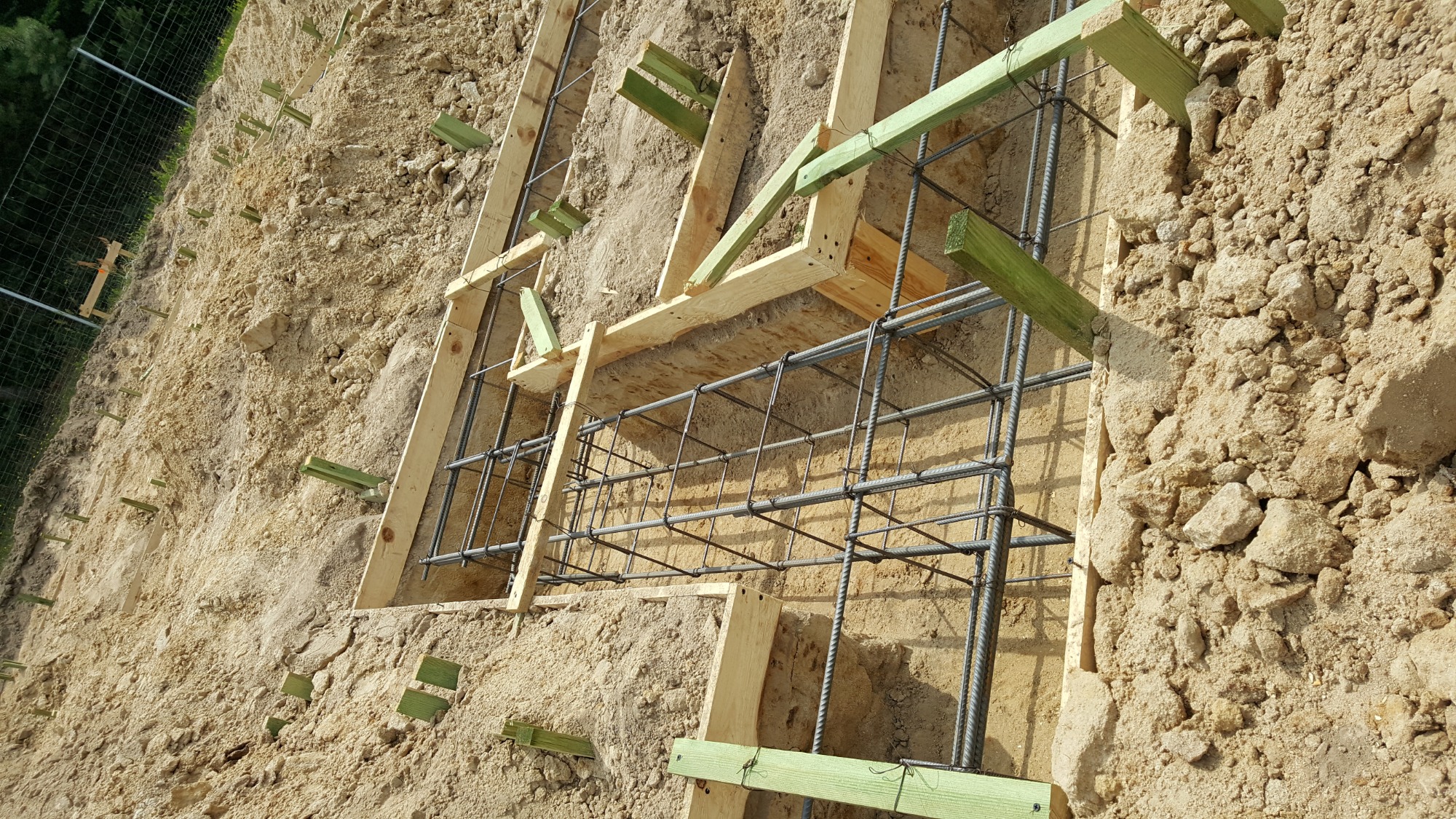 Zbrojenie fundamentu w wykopie, drewniane szalunki z zielonymi rozpórkami w piaszczystej ziemi.