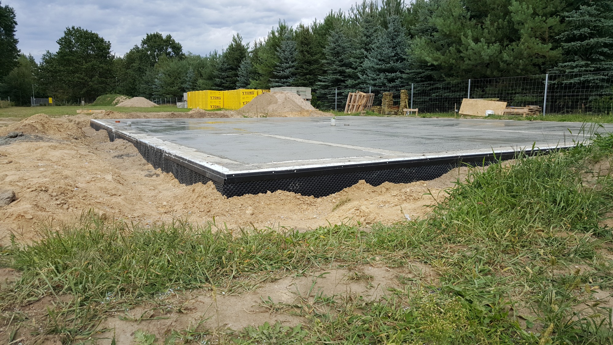 Wylany fundament z izolacją pionową, widoczne bloczki Ytong w tle, teren przygotowany pod budowę domu, trawa na pierwszym planie.