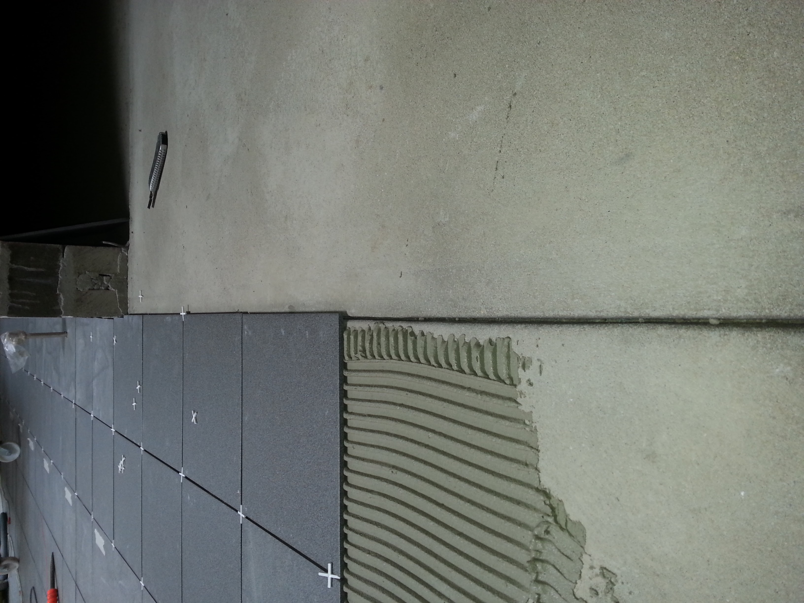 Układanie ciemnoszarych płytek na ścianie z użyciem krzyżyków dystansowych, widoczna warstwa kleju z charakterystycznym wzorem i nożyk do cięcia płytek.