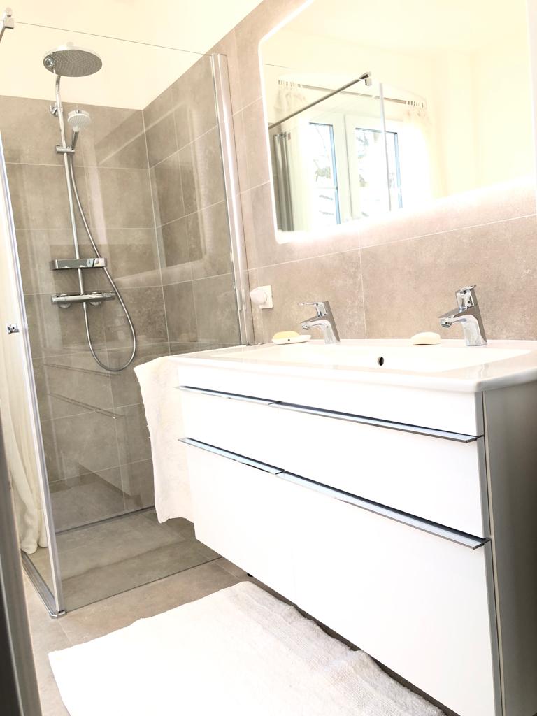 Nowoczesna łazienka z kabiną prysznicową typu walk-in, białą szafką podumywalkową z dwiema szufladami i podświetlanym lustrem, widok z boku na wejście do kabiny.