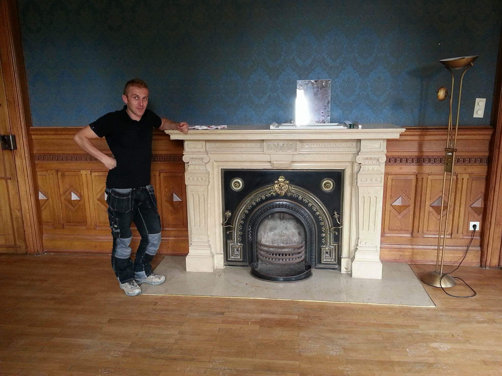Mężczyzna w roboczych spodniach opiera się o ozdobny, odnowiony kominek z czarnym wkładem w pokoju z drewnianą boazerią i wzorzystą, niebieską tapetą.
