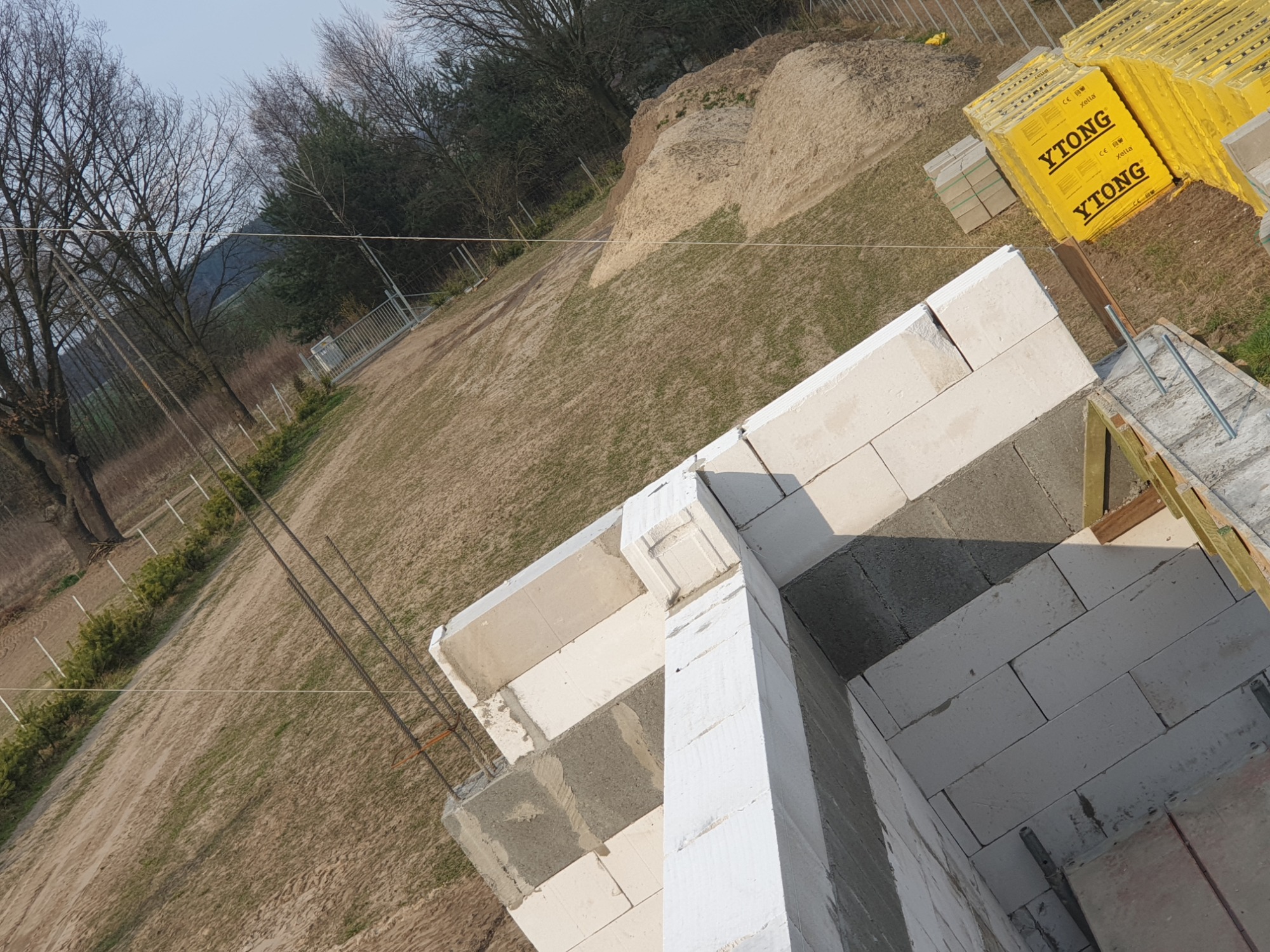 Wznoszone ściany domu z bloczków Ytong, widok z góry na narożnik budynku w trakcie budowy, w tle stos palet z bloczkami i hałda ziemi.