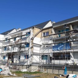 FIRMA USŁUGOWA PIOTR ZAROŚLIŃSKI - Wielorodzinny budynek w trakcie ocieplania elewacji, widoczne rusztowania, częściowo ocieplone ściany, materiały budowlane na trawniku i pracownicy w trakcie pracy.