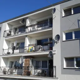 Odnowiony blok mieszkalny z balkonami i antenami satelitarnymi, częściowo zasłonięty rusztowaniem.