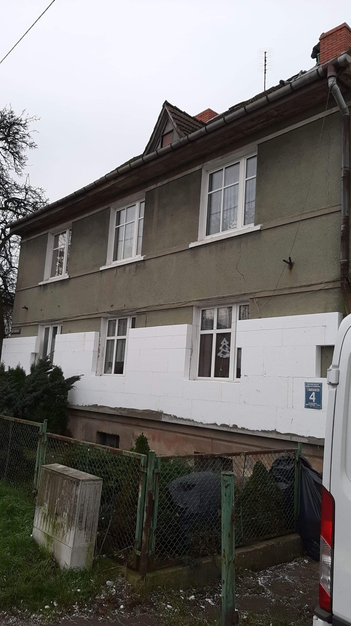 Dwupiętrowy dom z szarą elewacją w trakcie ocieplania styropianem, widoczne białe płyty izolacyjne na dolnej części fasady, z numerem domu '4' na ścianie.