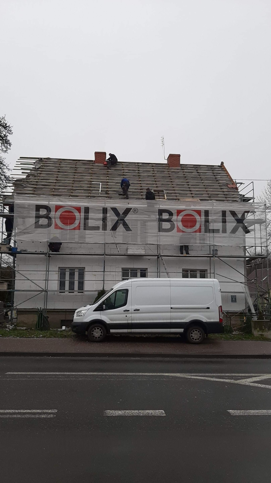 Dom w trakcie budowy, z widocznymi rusztowaniami, częściowo odsłoniętym dachem i reklamą firmy Bolix na elewacji, biały samochód dostawczy zaparkowany obok.