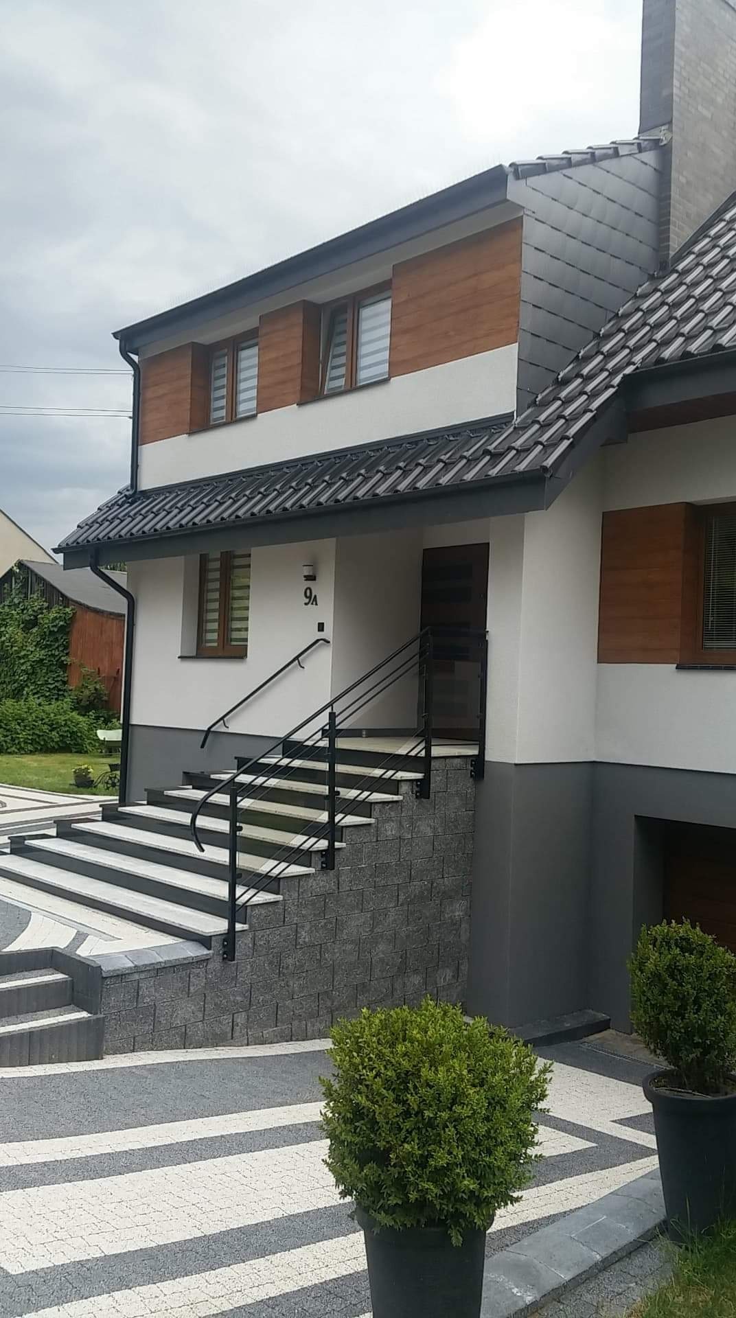 Dwukondygnacyjny dom z elewacją w kolorze białym i drewnianym wykończeniem, ciemna dachówka, schody wejściowe z szarych płyt z czarną balustradą, kostka brukowa w biało-szare pasy.