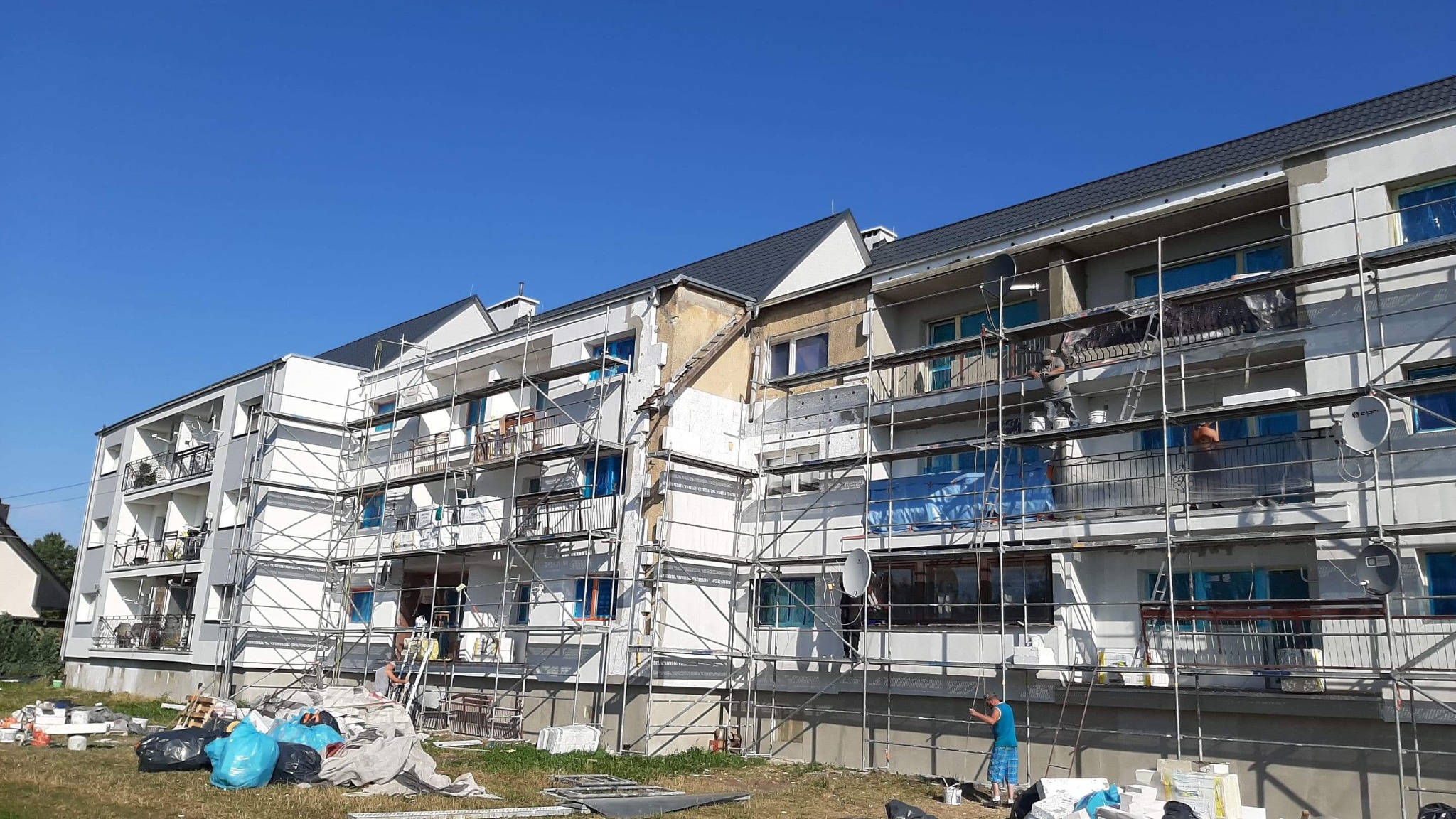 Wielorodzinny budynek w trakcie ocieplania elewacji, widoczne rusztowania, częściowo ocieplone ściany, materiały budowlane na trawniku i pracownicy w trakcie pracy.