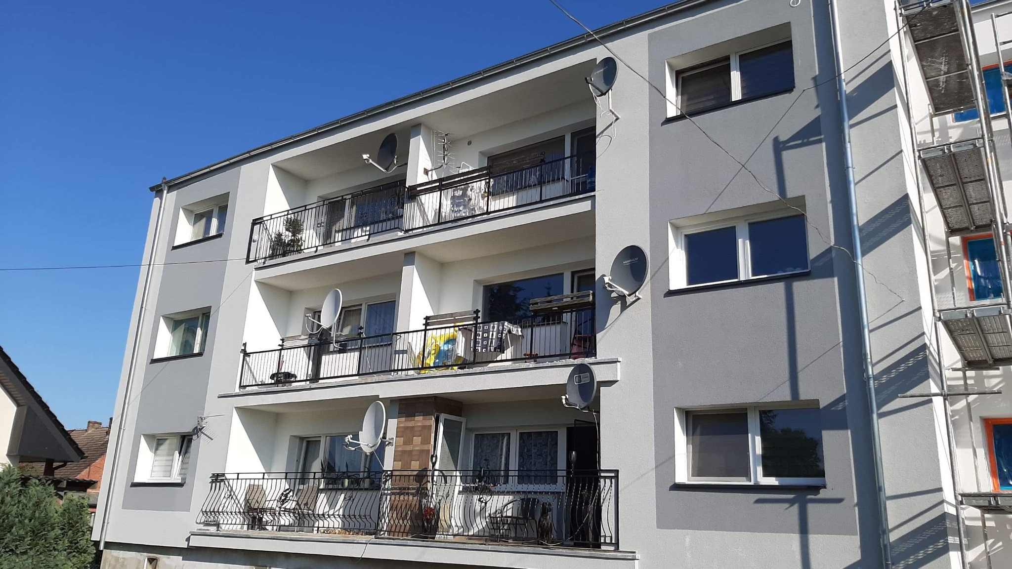 Odnowiony blok mieszkalny z balkonami i antenami satelitarnymi, częściowo zasłonięty rusztowaniem.
