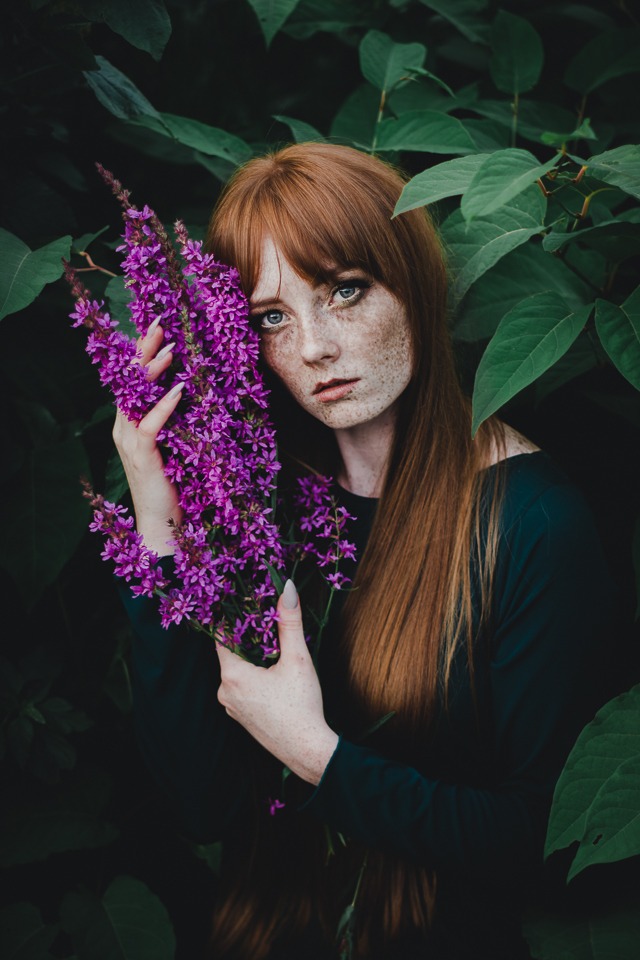 Portret rudej kobiety z piegami, trzymającej bukiet fioletowych kwiatów na tle zielonych liści.