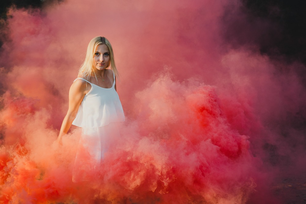 Kobieta w białej sukience pozuje w otoczeniu kłębów pomarańczowo-różowego dymu, patrząc w obiektyw.
