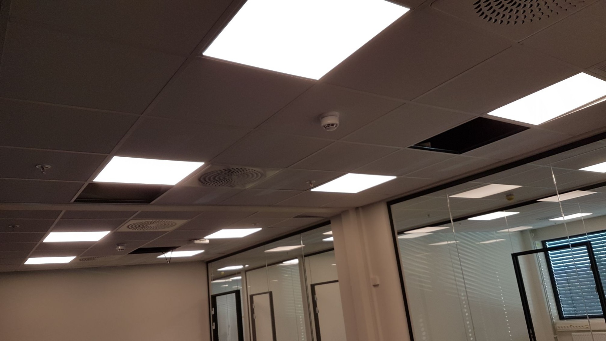Sufit podwieszany w biurze z panelami LED, kratkami wentylacyjnymi i czujnikiem dymu; widoczne braki w panelach.