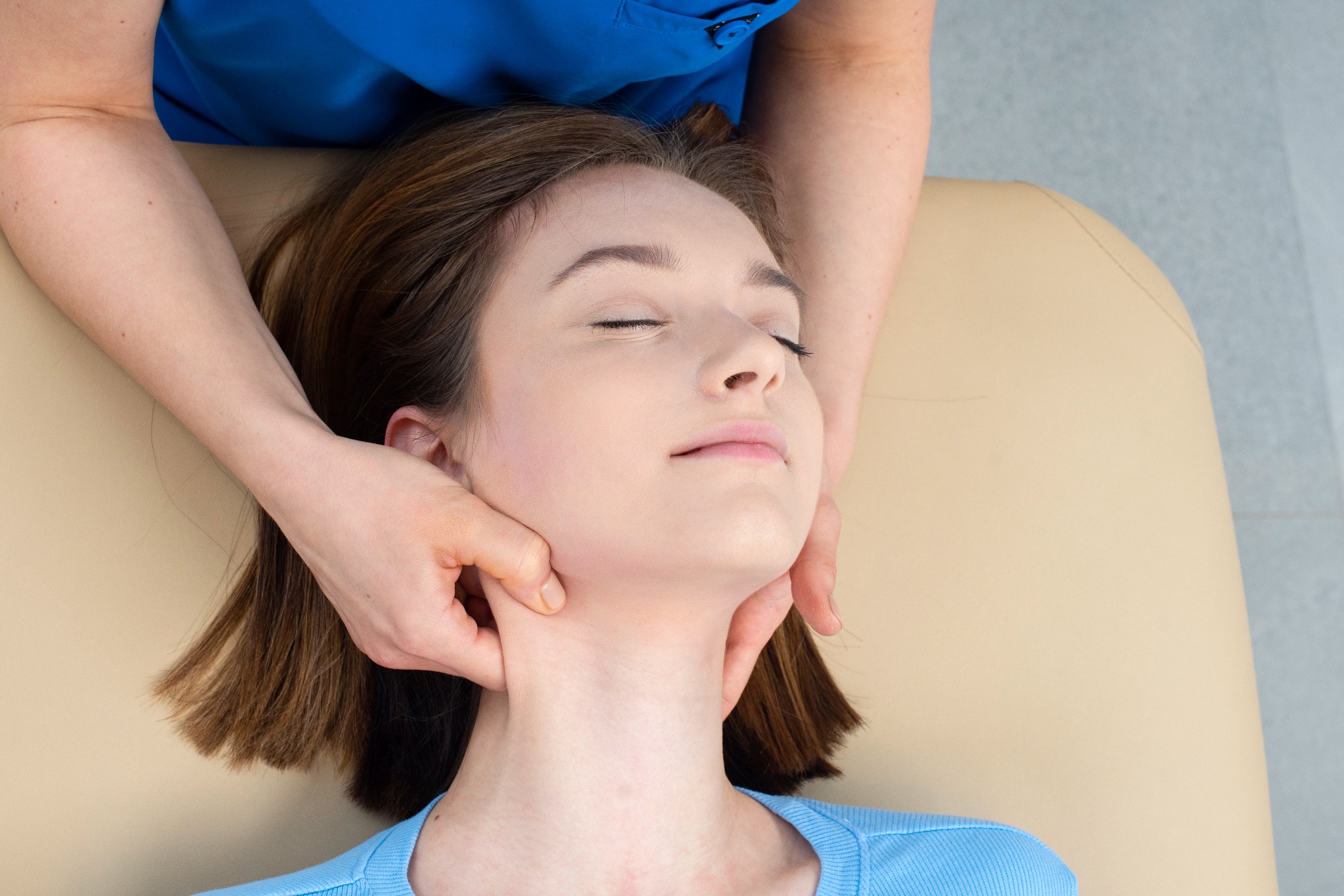 Masaż szyi wykonywany na młodej kobiecie leżącej na beżowym stole do masażu, z widocznymi dłońmi masażysty i fragmentem niebieskiego uniformu.