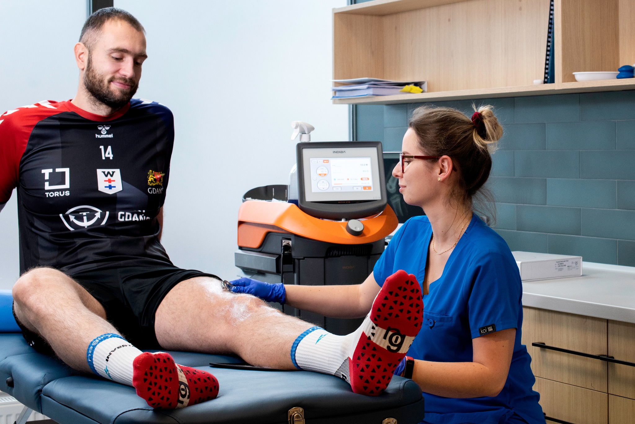 Fizjoterapeutka w niebieskim uniformie wykonuje zabieg Indiba na kolanie pacjenta w koszulce sportowej, widoczny ekran urządzenia Indiba, czerwone skarpety antypoślizgowe pacjenta.