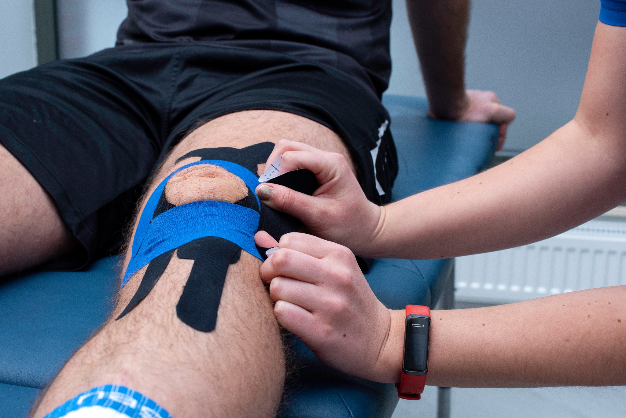 Aplikacja kinesiotapingu na kolano pacjenta w Gdańsku; widoczne dłonie terapeuty z czerwonym smartbandem i fragmenty taśm w kolorze niebieskim i czarnym.