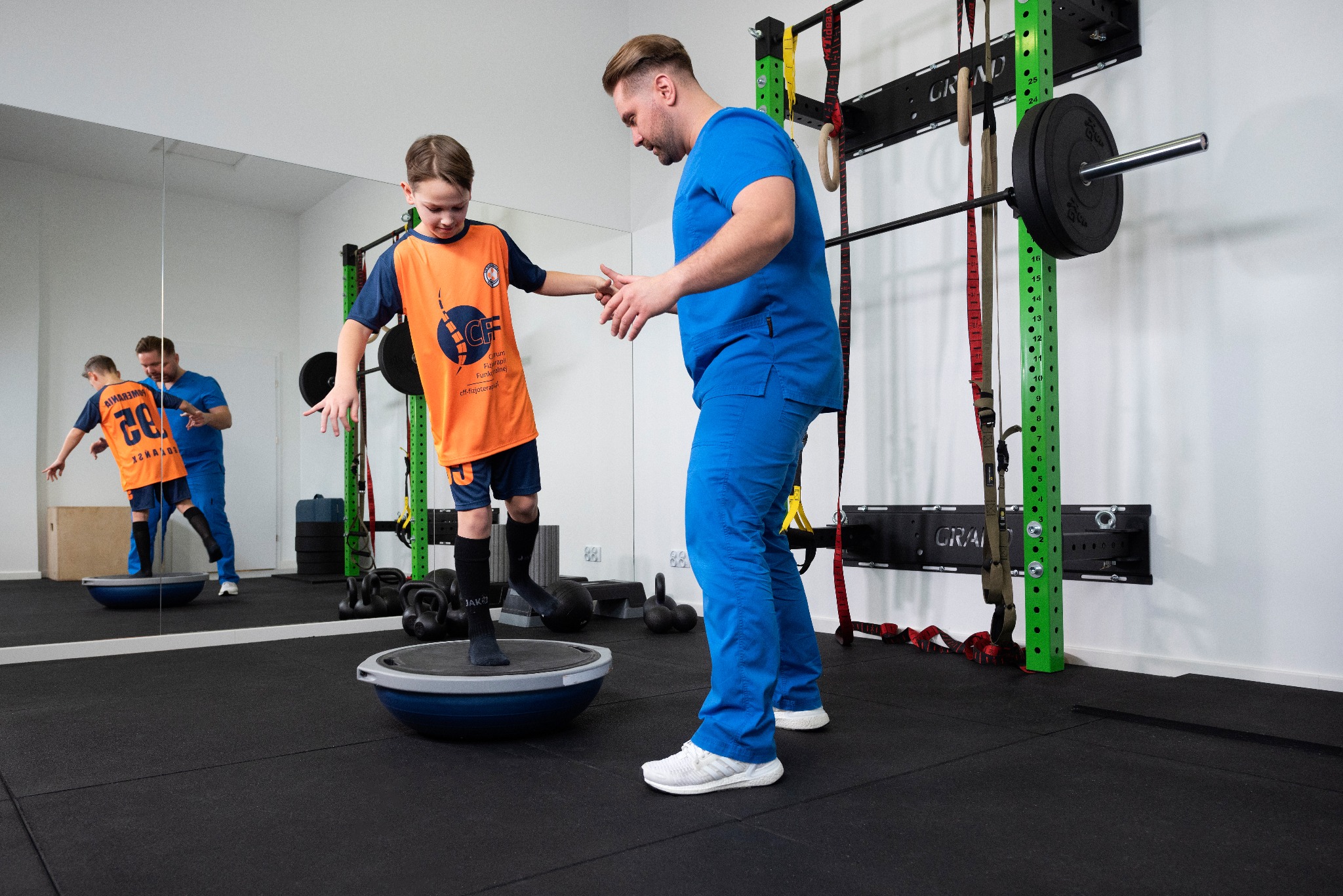 Chłopiec w sportowym stroju ćwiczy równowagę na platformie pod okiem terapeuty w niebieskim uniformie, widoczne lustro z odbiciem, drabinki gimnastyczne i obciążenia.