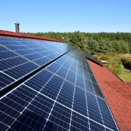 Panele słoneczne na czerwonym dachu domu jednorodzinnego, widok z góry, w tle las iglasty i błękitne niebo.