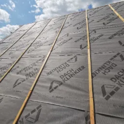 Montaż membrany dachowej z widocznym logo 'DACH KLINKIER' na drewnianej konstrukcji dachu pod kątem, z fragmentem błękitnego nieba z chmurami w tle.