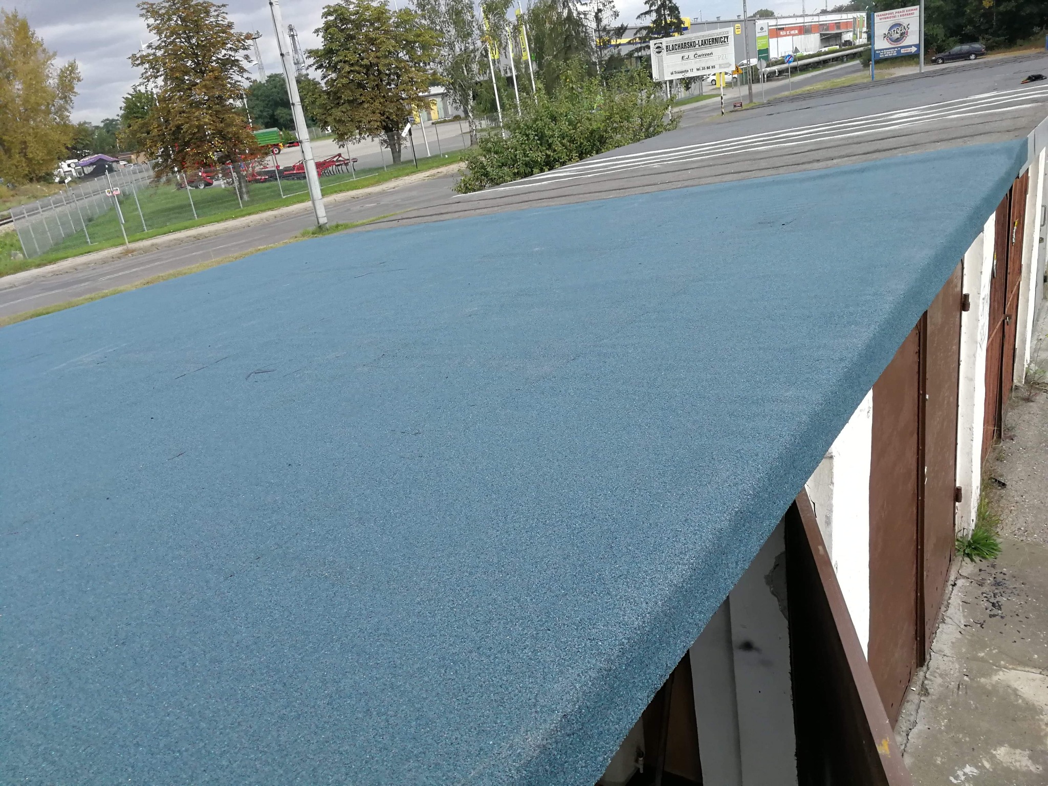 Niebieska, gumowa powłoka na płaskim dachu garażu z brązowymi drzwiami, widok z góry, w tle ulica i budynki.