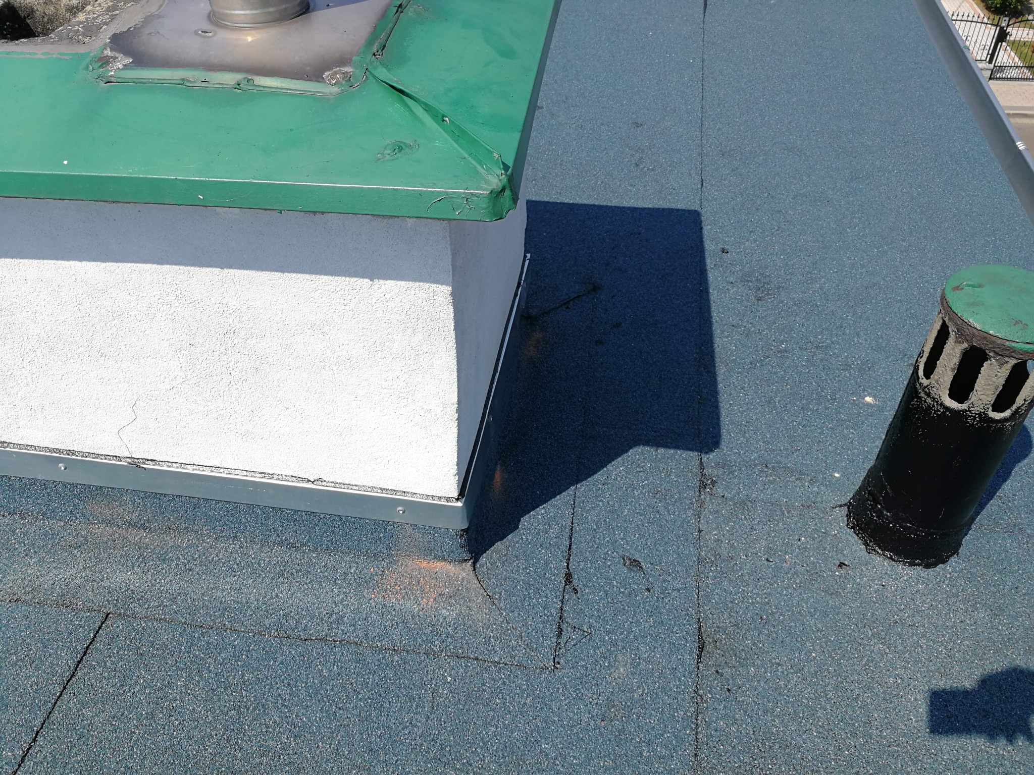 Widok z góry na fragment dachu pokrytego papą w kolorze niebiesko-szarym, z kominem wentylacyjnym i obróbką komina, widoczne ślady po naprawach.