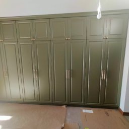 Duża, oliwkowa szafa wnękowa z frezowanymi frontami i złotymi uchwytami, zajmująca całą ścianę, na podłodze kartony zabezpieczające, widoczna żarówka.