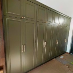 Długa, oliwkowa szafa wnękowa z frezowanymi drzwiami i złotymi uchwytami, sięgająca sufitu. Podłoga zabezpieczona kartonem.