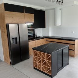 Nowoczesna kuchnia z wyspą: drewniane i czarne szafki, marmurowy blat, srebrna lodówka side-by-side, biały okap cylindryczny, szare krzesła.