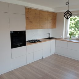 Nowoczesna kuchnia z białymi szafkami i drewnianymi akcentami. Czarny piekarnik w zabudowie, marmurowy backsplash i designerska lampa. Widok na okno z zielenią.