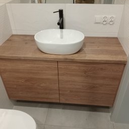 Wojciech Kozłowski - Drewniana szafka łazienkowa z owalną umywalką nablatową i czarną armaturą. Białe płytki z subtelnym wzorem na ścianie. Nowoczesny design.